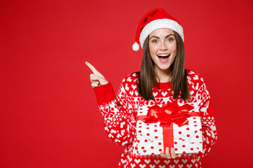 Excited young Santa woman in sweater Christmas hat hold present box with gift ribbon bow pointing index finger aside up isolated on red background. Happy New Year celebration merry holiday concept.