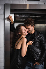 Smiling man with hand in pocket standing near cheerful brunette woman touching jacket, while leaning on elevator entrance