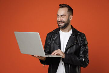 Smiling handsome attractive young bearded man 20s wearing basic white t-shirt black leather jacket standing working on laptop pc computer isolated on bright orange colour background studio portrait.