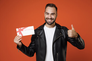 Smiling young bearded man wearing basic white t-shirt, black leather jacket standing hold gift certificate showing thumb up looking camera isolated on bright orange colour background studio portrait.