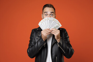 Shocked young bearded man in basic white t-shirt, black leather jacket standing covering face with fan of cash money in dollar banknotes looking camera isolated on orange background studio portrait.