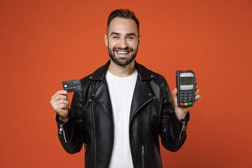 Smiling young man in white t-shirt, black leather jacket standing hold wireless modern bank payment terminal to process and acquire credit card payments isolated on orange background studio portrait.