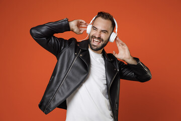 Laughing young bearded man wearing casual basic white t-shirt, black leather jacket standing listening music with headphones looking aside isolated on bright orange colour background studio portrait.