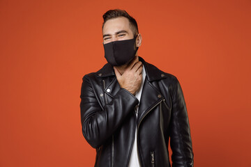 Obraz premium Sick young bearded man 20s in white t-shirt black leather jacket face mask safe from coronavirus virus covid-19 feeling sore throat, put hand on neck isolated on orange background studio portrait.