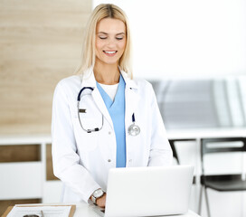 Blond female physician is lsmiling while using laptop computer. Woman-doctor at work in clinic excited and happy of her profession. Medicine concept