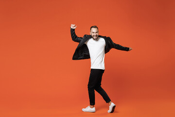 Fototapeta na wymiar Full length side view of laughing young bearded man in basic white t-shirt black leather jacket dancing turning around to camera spreading hands isolated on orange colour background studio portrait.
