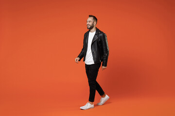 Fototapeta na wymiar Full length side view of smiling cheerful young bearded man 20s wearing basic white t-shirt black leather jacket standing walking going looking aside isolated on orange background studio portrait.