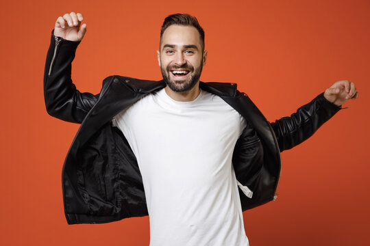 Cheerful laughing young bearded man 20s wearing basic white t-shirt, black leather jacket standing dancing spreading hands looking camera isolated on bright orange colour background, studio portrait.