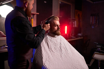 confident caucasian barber mster cuts hair and beard of men in the barbershop, professional...