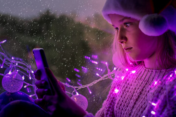 Cute teenage girl in Santa hat using mobile with falling snow outside the window during Christmas holidays