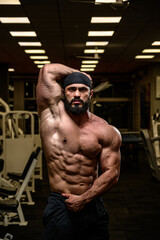 Plakat strong young athlete bearded male wearing bandana showing abdominal muscle in dark night gym