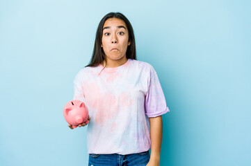 Young asian woman holding a pink bank over isolated background shrugs shoulders and open eyes...