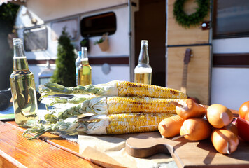 Sausages, corn and bottles of beer on wooden table near motorhome on sunny day. Camping season