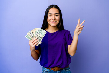 Young asian woman holding money isolated on purple background joyful and carefree showing a peace symbol with fingers.