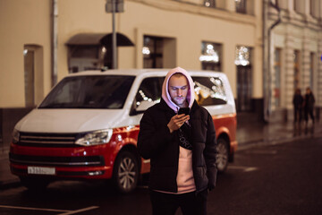 Fototapeta na wymiar Man in hood looking into his smartphone in background of car on night street.