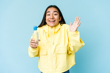 Young asian woman holding a bottle of water isolated on blue background receiving a pleasant...