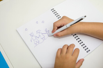 Top view of a child's hand draws a family with a pen on a white notebook.