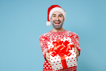 Cheerful Northern bearded man frozen snow face in Santa hat Christmas sweater hold present box with gift ribbon bow isolated on blue background. Happy New Year celebration holiday winter time concept.