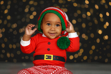 Cute little African American baby wearing elf hat against blurred lights on dark background. Christmas celebration