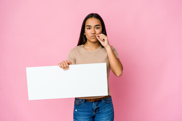 Fototapeta na wymiar Young asian woman holding a blank paper for white something over isolated background with fingers on lips keeping a secret.