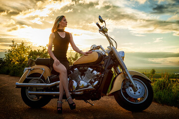 Fototapeta na wymiar Attractive female brunette motorcyclist with motorcycle in a summer evening during sunset. Beautiful girl on nature and sky with sun and clouds background. Adventure concept