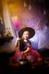 Little cute blonde girl looking as witch in special dress and hat in room decorated for Halloween. Witchcraft and wizardry in carnival. Halloween style photo shoot.