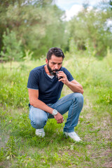 Naklejka na ściany i meble Stylish bearded man is resting in nature and vaping and letting off steam from an electronic cigarette. Non-tobacco smoking.