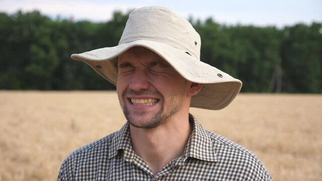 Portrait of happy smiling agronomist in hat looking into camera against the blurred background of wheat field. Young laughing farmer standing in the barley meadow. Concept of agricultural business