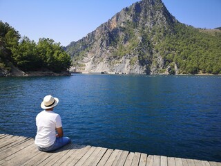 A man dressed in white, sat on the pier, staring into the deep blue ocean, where a mountain is reaching for the sky. Surrounded by a lush green forest and a perfectly clear sky.