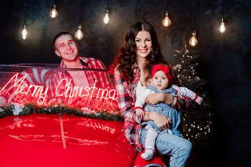 Young beautiful parents holding their little cute daughter in their arms having fun near retro car in studio. Christmas family look. New Year's scenery.