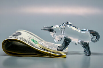 Figurine of a bull with banknotes on a gray background. Business concept, stock exchange, finance. Selective focus. The US dollars are stacked on top of each other, with a glass bull above it.