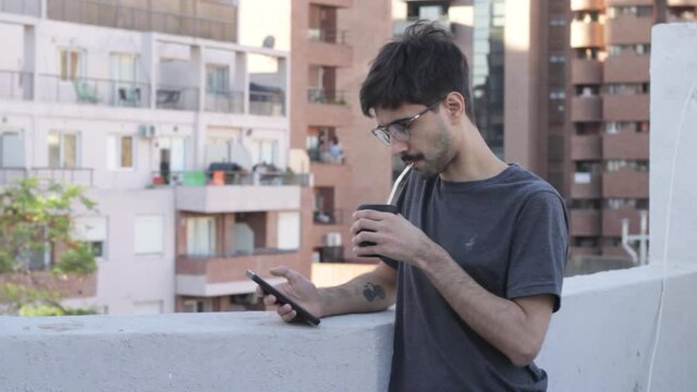 Young handsome man using smartphone and drinking mate in a rooftop with city background