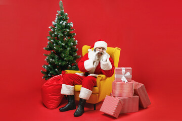 Santa Claus man in Christmas hat suit coat sit in armchair with fir tree presents gifts boxes screaming with hands gesture near mouth isolated on red background. New Year celebration holiday concept.