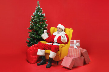 Excited Santa Claus man in Christmas hat suit coat sit in armchair with fir tree present gifts doing winner gesture isolated on red background studio. Happy New Year celebration merry holiday concept.