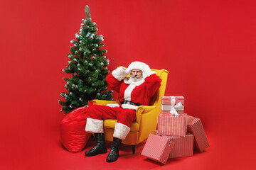 Displeased Santa Claus man in Christmas hat suit sit in armchair with fir tree presents gifts put hands on head isolated on red background studio. Happy New Year celebration merry holiday concept.