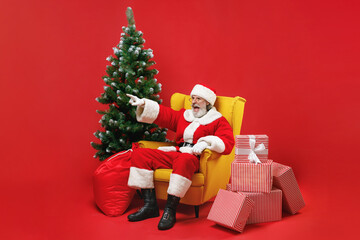 Angry Santa Claus man in Christmas hat suit sit in armchair with fir tree presents gifts point index finger aside swearing isolated on red background. Happy New Year celebration merry holiday concept.