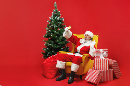 Funny Santa Claus man in Christmas suit sit in armchair with fir tree gifts doing selfie shot on mobile phone greeting with hand isolated on red background. Happy New Year celebration holiday concept.