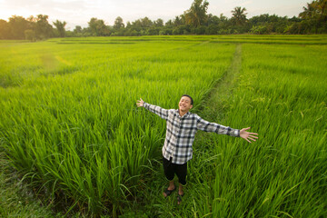 Obraz na płótnie Canvas Good mood of young asian man breathing fresh air and rised arms up in middle green fields at sunset background. Environment friendly, leisure during travel at rural country concept.