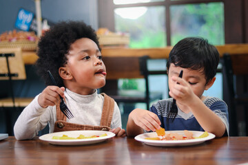 Cute African American boy with curly and adorable Asian kid eating meal at the table indoor, happy children having food in a restaurant, enjoy their meal together