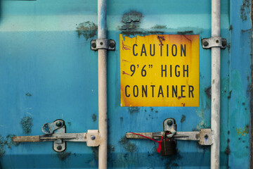 The back door of blue heavy-duty container with locks and sealed padlock. Container is very rusty and aged. Yellow warning sign about high container height is marked on the door
