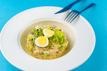 Traditional Russian salad Olivier. Christmas and New year food. White plat with fork and knife on blue background with copy space. Top view, flat lay. Microgreen, quail eggs. Serving at a restaurant.