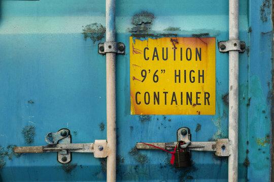 The Back Door Of Blue Heavy-duty Container With Locks And Sealed Padlock. Container Is Very Rusty And Aged. Yellow Warning Sign About High Container Height Is Marked On The Door