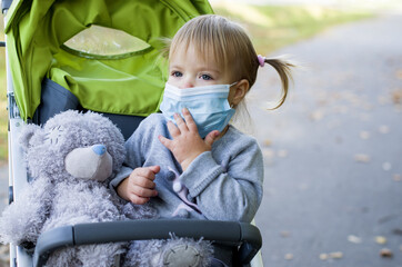 Сute little girl wearing protective surgical face mask sitting in stroller during coronavirus or...