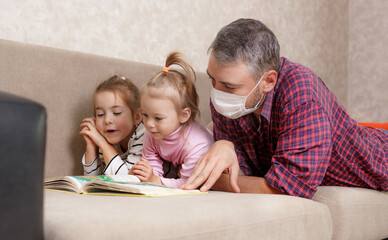 Fototapeta premium A young father at home in self-isolation reads a book to his beloved daughters.