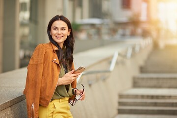 Side view of a young and happy cheerful business woman using her smartphone and smiling, looking aside while standing against blurred urban background outdoors