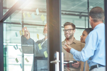 Group of creative coworkers putting colorful sticky notes on a glass wall, discussing something and smiling while standing in the modern office