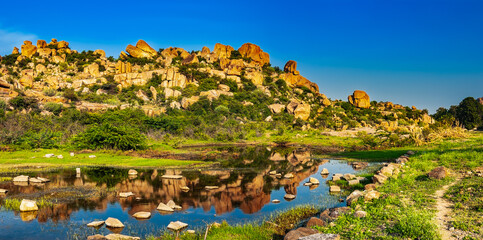 Tropical exotic landscape with ancient big rocks and stones, path by bright blue lake under blue sky in Hampi, Karnataka, India. Panoramic view of beautiful valley, stunning Indian nature scenery