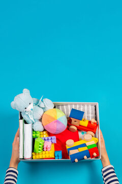 Donation Concept. Kid Hands With Donate Box With Clothes, Books And Toys On Light Blue Background. Top View