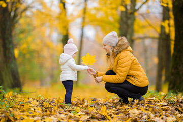 Young adult mother giving big bright yellow maple leaf to little daughter at park. Golden autumn....