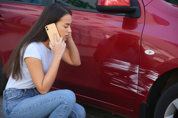 Stressed woman talking on phone near car with scratch outdoors